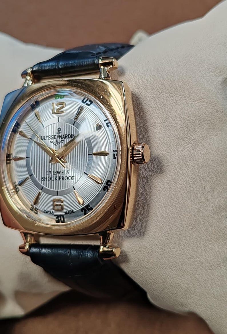 May include: A gold-tone Ulysse Nardin wristwatch with a square case and a black leather strap. The silver dial features a striped pattern, gold-tone hands, gold-tone hour markers, and the text Ulysse Nardin, 17 Jewels, Shock Proof, and Swiss Made.