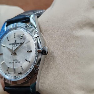 May include: A silver-toned Ulysse Nardin wristwatch with a black leather band. The watch face is silver with black hands and hour markers. The watch reads "17 Jewels Shock Proof" and "Swiss Made". The watch is on a wrist.