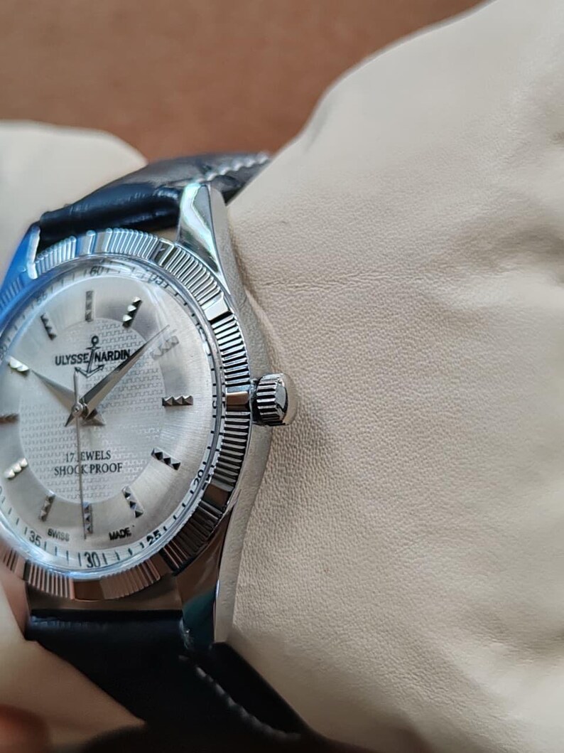 May include: A silver-toned Ulysse Nardin wristwatch with a black leather band. The watch face is silver with black hands and hour markers. The watch reads "17 Jewels Shock Proof" and "Swiss Made". The watch is on a wrist.