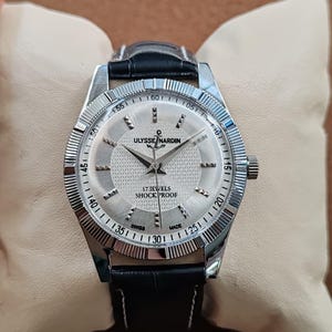 May include: A silver Ulysse Nardin watch with a black leather band. The watch face is silver with black numerals and hands. The text "17 JEWELS SHOCK PROOF" is visible. The watch is displayed on a cream-colored pillow.