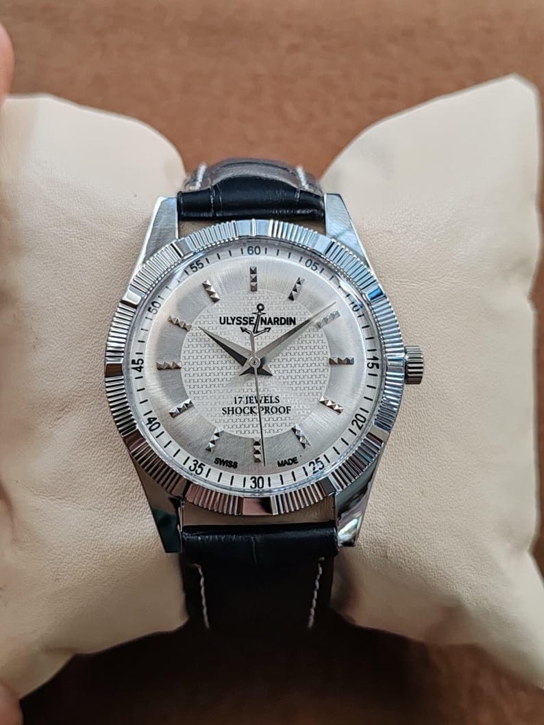 May include: A silver Ulysse Nardin watch with a black leather band. The watch face is silver with black numerals and hands. The text "17 JEWELS SHOCK PROOF" is visible. The watch is displayed on a cream-colored pillow.