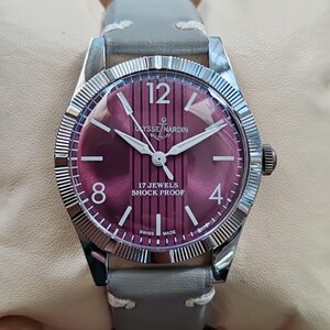 May include: A wristwatch with a circular silver-toned case and a fluted bezel. The dial is a deep magenta color with vertical stripes, featuring the text Ulysse Nardin, 17 Jewels, Shock Proof, and Swiss Made. It has a grey leather strap.