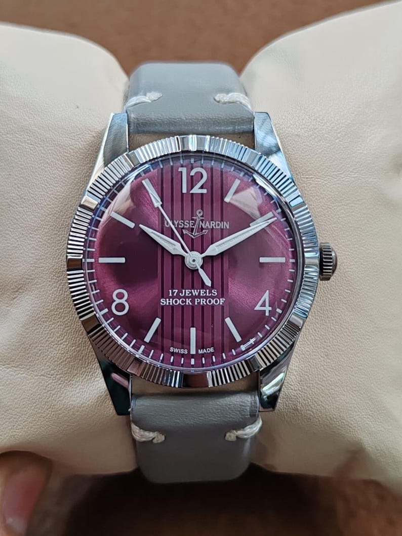 May include: A wristwatch with a circular silver-toned case and a fluted bezel. The dial is a deep magenta color with vertical stripes, featuring the text Ulysse Nardin, 17 Jewels, Shock Proof, and Swiss Made. It has a grey leather strap.
