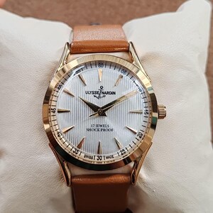 May include: A gold-tone wristwatch with a white, vertically textured dial featuring gold-tone hands and hour markers. The dial displays the text ULYSSE NARDIN, an anchor logo, 17 JEWELS, SHOCK PROOF, and SWISS MADE. It has a tan leather strap.