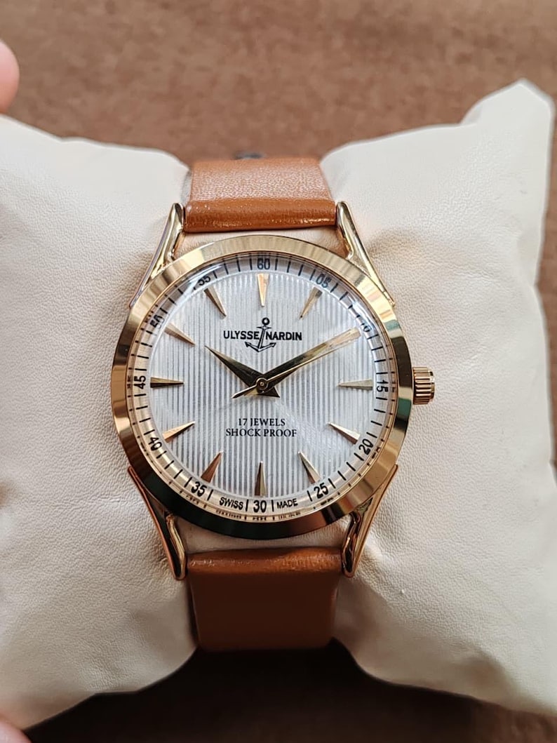 May include: A gold-tone wristwatch with a white, vertically textured dial featuring gold-tone hands and hour markers. The dial displays the text ULYSSE NARDIN, an anchor logo, 17 JEWELS, SHOCK PROOF, and SWISS MADE. It has a tan leather strap.