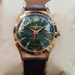 May include: A gold-tone wristwatch with a dark green dial featuring vertical pinstripes, gold-tone indices, and the text Ulysse Nardin, 17 Jewels, Shock Proof, and Swiss Made. The watch has a red second hand and a brown leather strap.