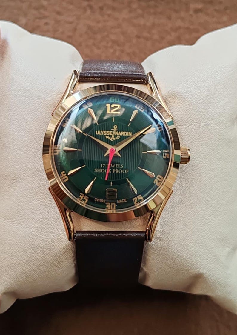 May include: A gold-tone wristwatch with a dark green dial featuring vertical pinstripes, gold-tone indices, and the text Ulysse Nardin, 17 Jewels, Shock Proof, and Swiss Made. The watch has a red second hand and a brown leather strap.