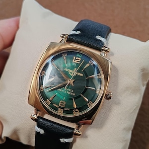 K&ouml;nnte beinhalten: Eine goldfarbene quadratische Armbanduhr mit gr&uuml;nem Sonnenschliff-Zifferblatt, goldenen Indizes, arabischen Ziffern auf 12 und 6 sowie der Aufschrift Ulysse Nardin, 17 Jewels, Shock Proof und Swiss Made. Sie hat ein schwarzes Lederarmband mit wei&szlig;en N&auml;hten.
