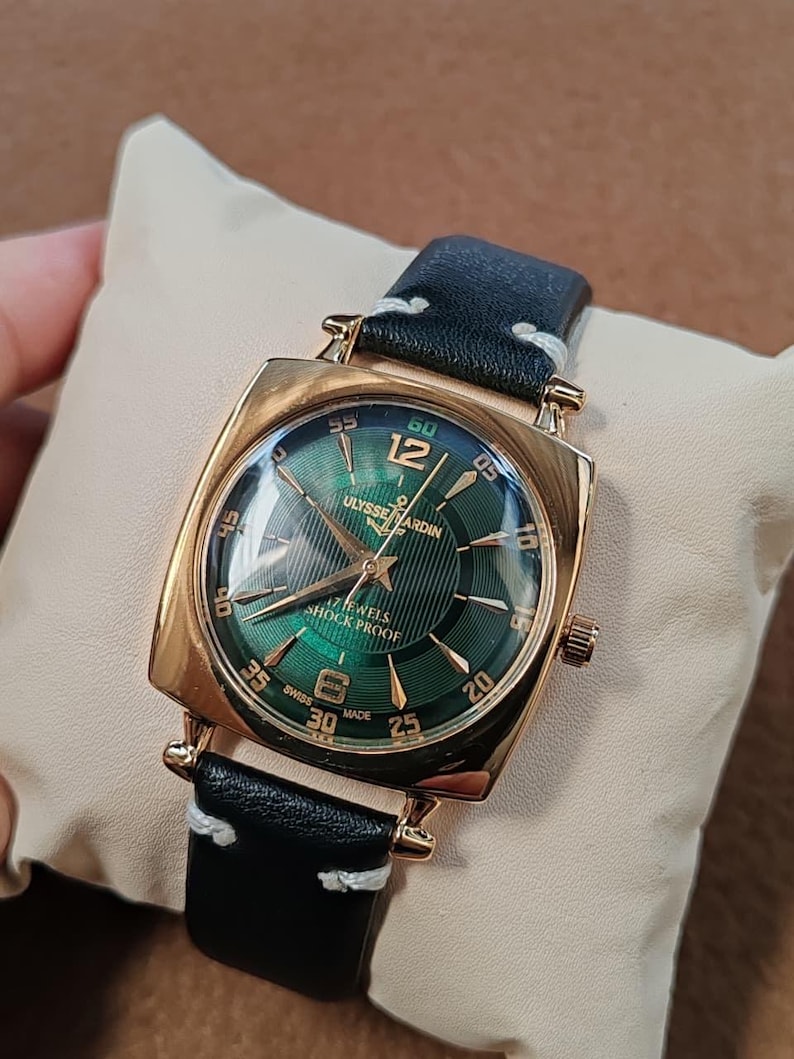 K&ouml;nnte beinhalten: Eine goldfarbene quadratische Armbanduhr mit gr&uuml;nem Sonnenschliff-Zifferblatt, goldenen Indizes, arabischen Ziffern auf 12 und 6 sowie der Aufschrift Ulysse Nardin, 17 Jewels, Shock Proof und Swiss Made. Sie hat ein schwarzes Lederarmband mit wei&szlig;en N&auml;hten.