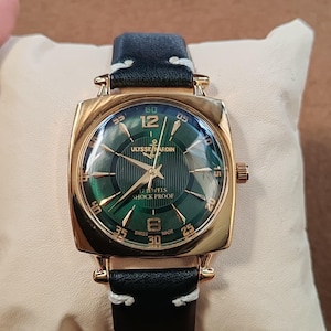 K&ouml;nnte beinhalten: Eine goldfarbene Armbanduhr mit quadratischem Geh&auml;use und einem gr&uuml;nen Zifferblatt mit vertikalen Streifen. Das Zifferblatt zeigt den Text ULYSSE NARDIN, 17 JEWELS, SHOCK PROOF und SWISS MADE. Sie hat ein schwarzes Lederarmband mit wei&szlig;en N&auml;hten.