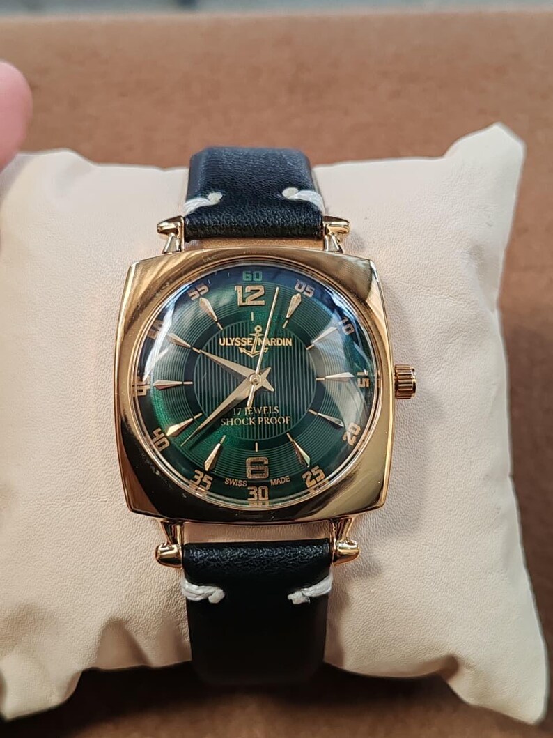 K&ouml;nnte beinhalten: Eine goldfarbene Armbanduhr mit quadratischem Geh&auml;use und einem gr&uuml;nen Zifferblatt mit vertikalen Streifen. Das Zifferblatt zeigt den Text ULYSSE NARDIN, 17 JEWELS, SHOCK PROOF und SWISS MADE. Sie hat ein schwarzes Lederarmband mit wei&szlig;en N&auml;hten.