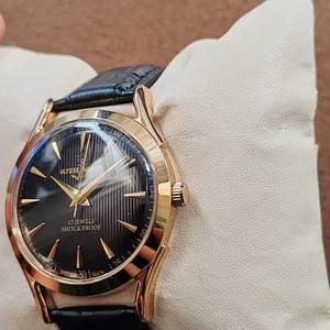 Puede incluir: Un reloj de pulsera dorado con esfera negra texturizada, manecillas doradas e &iacute;ndices dorados. La esfera muestra el texto Ulysse Nardin, 17 Jewels, Shock Proof y Swiss Made. Cuenta con una correa de cuero negro y caja redonda dorada.