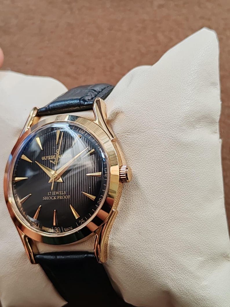 Puede incluir: Un reloj de pulsera dorado con esfera negra texturizada, manecillas doradas e &iacute;ndices dorados. La esfera muestra el texto Ulysse Nardin, 17 Jewels, Shock Proof y Swiss Made. Cuenta con una correa de cuero negro y caja redonda dorada.