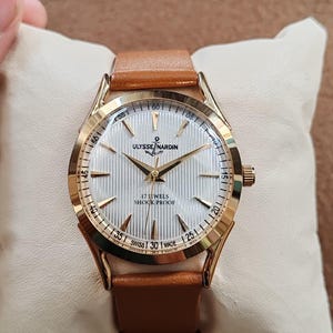 May include: A gold-tone wristwatch with a white, vertically textured dial, gold-tone hands, and hour markers. The dial features the text ULYSSE NARDIN, 17 JEWELS, SHOCK PROOF, and SWISS MADE. The watch has a tan leather strap and a round case.