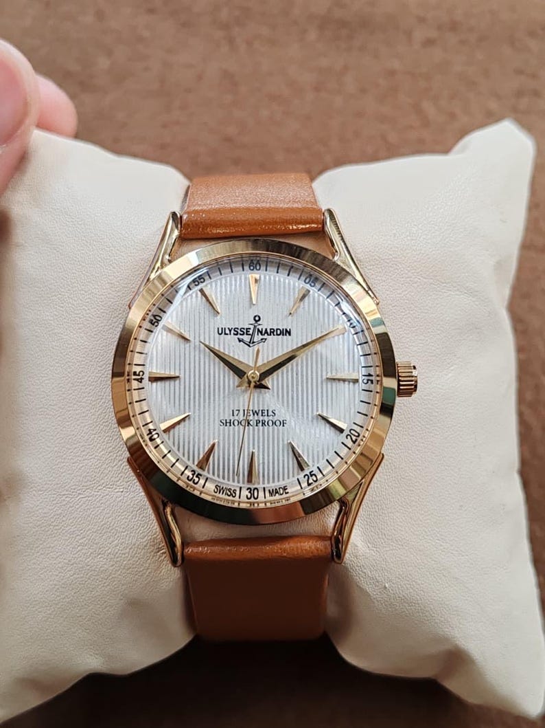 May include: A gold-tone wristwatch with a white, vertically textured dial, gold-tone hands, and hour markers. The dial features the text ULYSSE NARDIN, 17 JEWELS, SHOCK PROOF, and SWISS MADE. The watch has a tan leather strap and a round case.