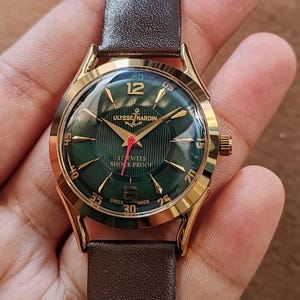 May include: A gold-tone wristwatch with a dark green dial featuring a textured pattern, gold-tone hour markers, and the text Ulysse Nardin, 17 Jewels, Shock Proof, and Swiss Made. The watch has a red second hand and a dark brown leather strap.