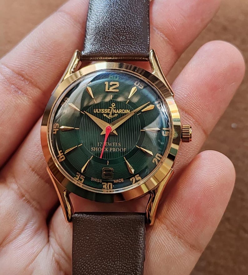 May include: A gold-tone wristwatch with a dark green dial featuring a textured pattern, gold-tone hour markers, and the text Ulysse Nardin, 17 Jewels, Shock Proof, and Swiss Made. The watch has a red second hand and a dark brown leather strap.