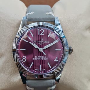 May include: A wristwatch with a silver-toned fluted bezel and a deep burgundy dial featuring vertical stripes. The dial displays the text Ulysse Nardin, 17 Jewels, Shock Proof, and Swiss Made, with white numerals at 12, 8, and 4 o'clock and a grey leather strap.