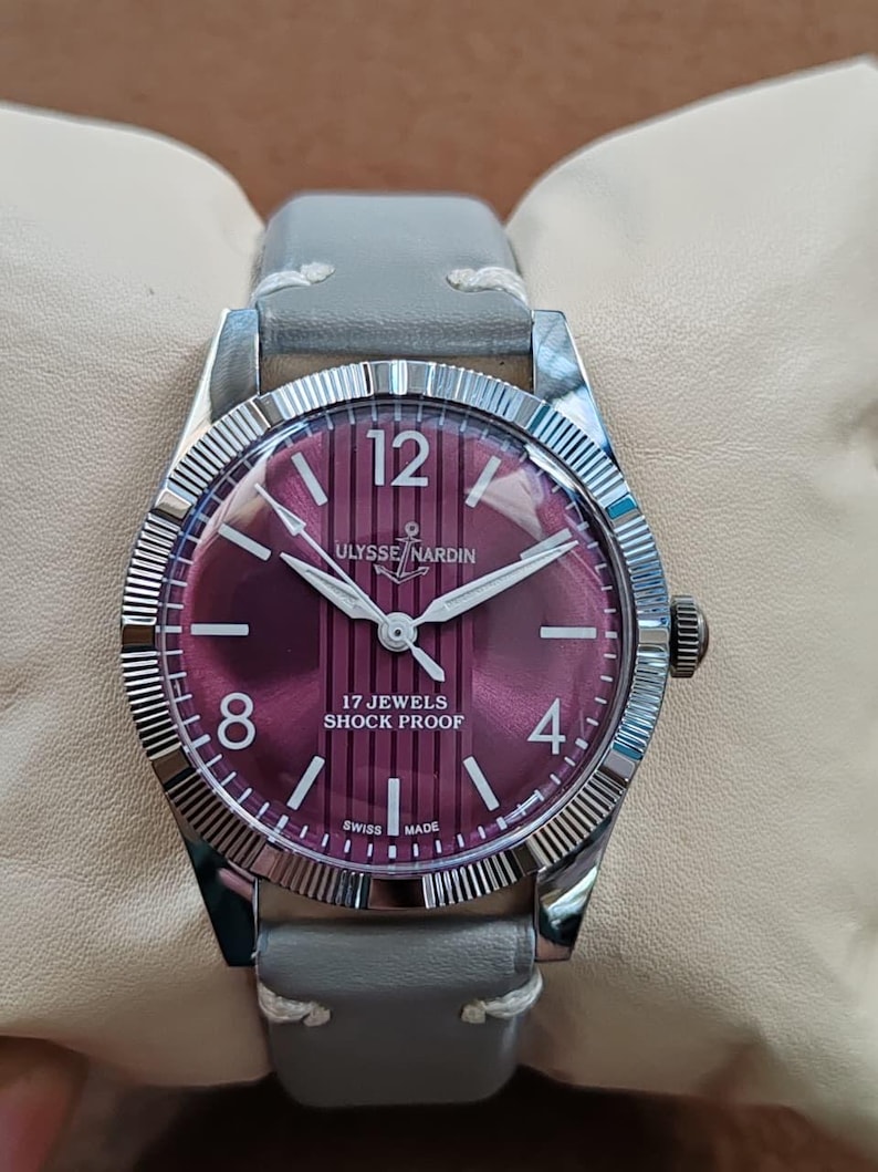 May include: A wristwatch with a silver-toned fluted bezel and a deep burgundy dial featuring vertical stripes. The dial displays the text Ulysse Nardin, 17 Jewels, Shock Proof, and Swiss Made, with white numerals at 12, 8, and 4 o'clock and a grey leather strap.