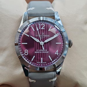 May include: A wristwatch with a round silver-toned case and a fluted bezel. The dial is deep purple with vertical stripes, featuring the text ULYSSE NARDIN, 17 JEWELS, SHOCK PROOF, and SWISS MADE. It has white hour markers, numerals 12, 8, 4, and a grey leather strap.