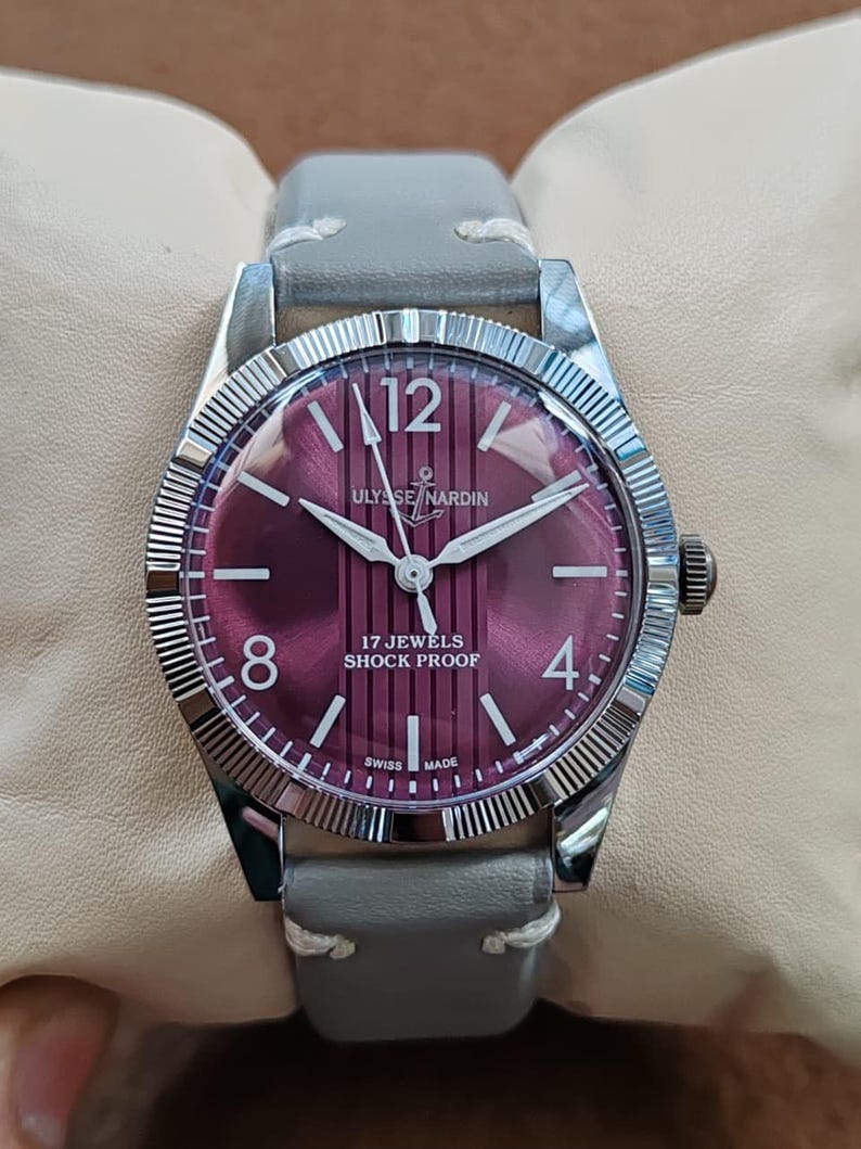 May include: A wristwatch with a round silver-toned case and a fluted bezel. The dial is deep purple with vertical stripes, featuring the text ULYSSE NARDIN, 17 JEWELS, SHOCK PROOF, and SWISS MADE. It has white hour markers, numerals 12, 8, 4, and a grey leather strap.
