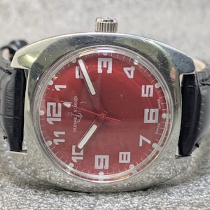 May include: A wristwatch with a polished silver-tone metal case and a vibrant red dial. The face features large white Arabic numerals, white luminous hands, and a black leather strap. The text Ulysse Nardin and Swiss Made are visible on the dial.