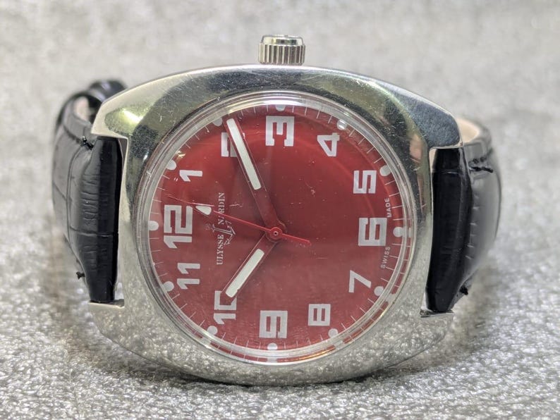 May include: A wristwatch with a polished silver-tone metal case and a vibrant red dial. The face features large white Arabic numerals, white luminous hands, and a black leather strap. The text Ulysse Nardin and Swiss Made are visible on the dial.