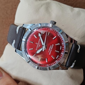 May include: A wristwatch with a vibrant red dial, silver-tone hands, and hour markers. The watch features a fluted silver bezel, a dark brown leather strap with white stitching, and text on the dial reading Ulysse Nardin, 17 Jewels, Shock Proof, Swiss Made.