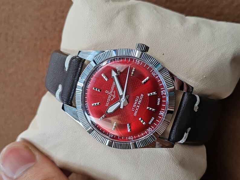 May include: A wristwatch with a vibrant red dial, silver-tone hands, and hour markers. The watch features a fluted silver bezel, a dark brown leather strap with white stitching, and text on the dial reading Ulysse Nardin, 17 Jewels, Shock Proof, Swiss Made.