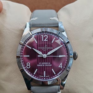 May include: A wristwatch with a circular silver-tone case and a fluted bezel. The dial is a deep magenta color with vertical stripes, featuring the text ULYSSE NARDIN, 17 JEWELS, SHOCK PROOF, and SWISS MADE. It has a grey leather strap with white stitching.