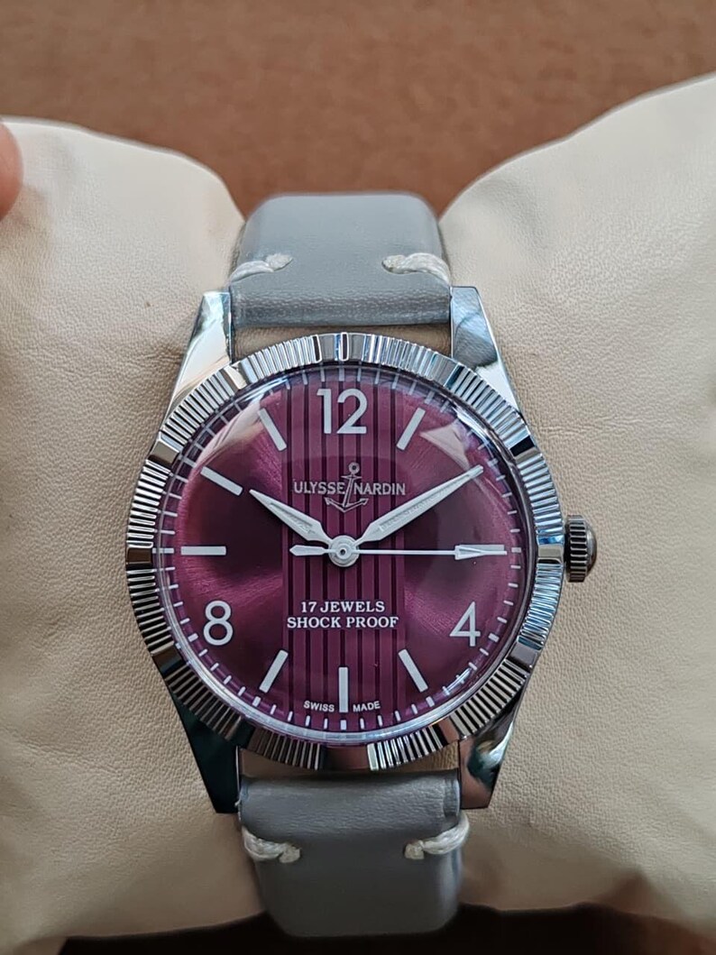 May include: A wristwatch with a circular silver-tone case and a fluted bezel. The dial is a deep magenta color with vertical stripes, featuring the text ULYSSE NARDIN, 17 JEWELS, SHOCK PROOF, and SWISS MADE. It has a grey leather strap with white stitching.