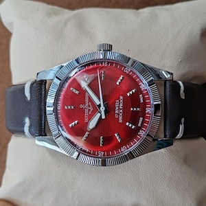 May include: A wristwatch with a circular red dial, silver-tone hands, and silver hour markers. The watch features a silver-tone fluted bezel, a dark brown leather strap with white stitching, and the text Ulysse Nardin, 17 Jewels, Shock Proof, Swiss Made.