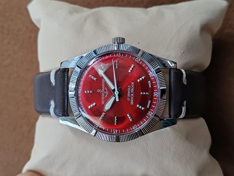 May include: A wristwatch with a circular red dial, silver-tone hands, and silver hour markers. The watch features a silver-tone fluted bezel, a dark brown leather strap with white stitching, and the text Ulysse Nardin, 17 Jewels, Shock Proof, Swiss Made.