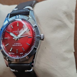 May include: A wristwatch with a circular silver-tone case and a vibrant red dial featuring silver-tone hour markers and hands. The dial displays the text Ulysse Nardin with an anchor logo, 17 Jewels, Shock Proof, and Swiss Made. It has a dark brown leather strap.