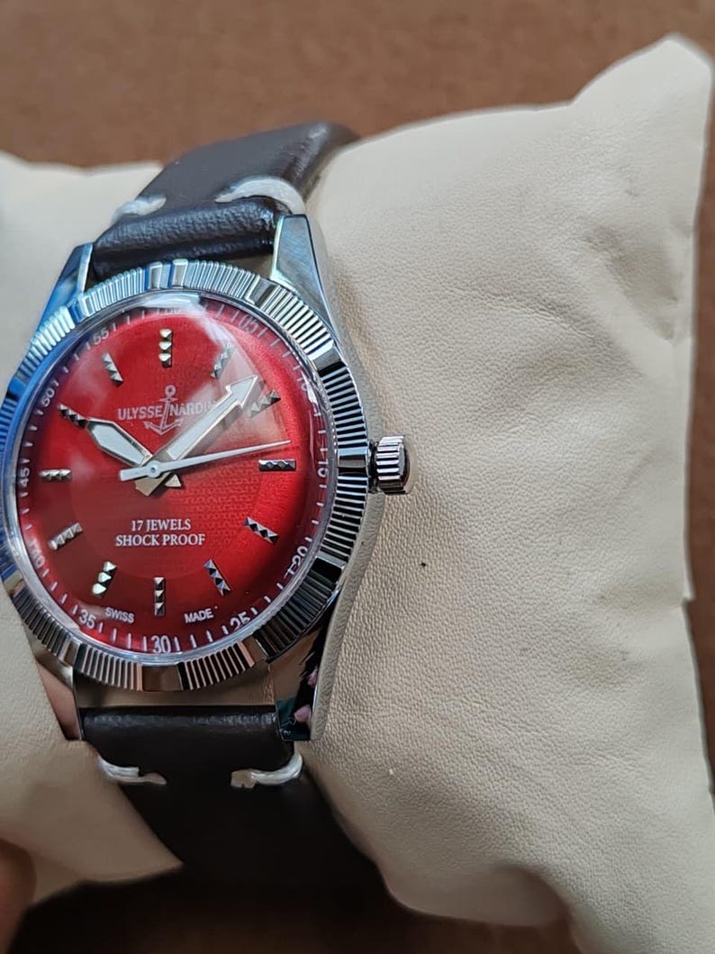May include: A wristwatch with a circular silver-tone case and a vibrant red dial featuring silver-tone hour markers and hands. The dial displays the text Ulysse Nardin with an anchor logo, 17 Jewels, Shock Proof, and Swiss Made. It has a dark brown leather strap.