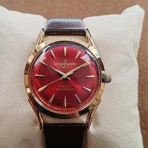 May include: A gold-tone Ulysse Nardin wristwatch featuring a vibrant red dial with a textured vertical striped pattern, gold-tone hour markers, and gold hands. The dial displays the text Ulysse Nardin, 17 Jewels, Shock Proof, and Swiss Made.