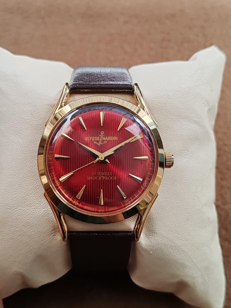 May include: A gold-tone Ulysse Nardin wristwatch featuring a vibrant red dial with a textured vertical striped pattern, gold-tone hour markers, and gold hands. The dial displays the text Ulysse Nardin, 17 Jewels, Shock Proof, and Swiss Made.