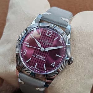 May include: A wristwatch with a round silver-tone fluted bezel and a deep magenta dial featuring vertical stripes. The dial displays the text Ulysse Nardin, 17 Jewels, Shock Proof, and Swiss Made, with silver hands and a grey leather strap.