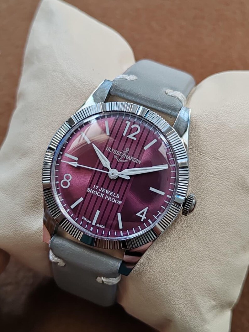 May include: A wristwatch with a round silver-tone fluted bezel and a deep magenta dial featuring vertical stripes. The dial displays the text Ulysse Nardin, 17 Jewels, Shock Proof, and Swiss Made, with silver hands and a grey leather strap.