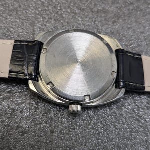 May include: A silver-toned wristwatch case featuring a circular back with a radial brushed finish and six notches along the perimeter. The watch includes a black leather strap with white stitching and a small metallic crown on the side of the case.