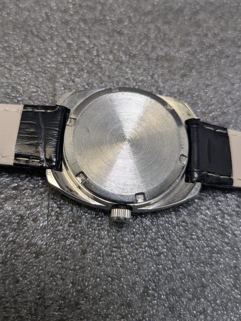 May include: A silver-toned wristwatch case featuring a circular back with a radial brushed finish and six notches along the perimeter. The watch includes a black leather strap with white stitching and a small metallic crown on the side of the case.
