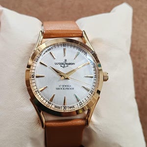May include: A gold-tone wristwatch with a tan leather strap and a white dial featuring vertical pinstripes. The dial displays the text Ulysse Nardin with an anchor logo, 17 Jewels, Shock Proof, and Swiss Made, with gold-tone hands and markers.