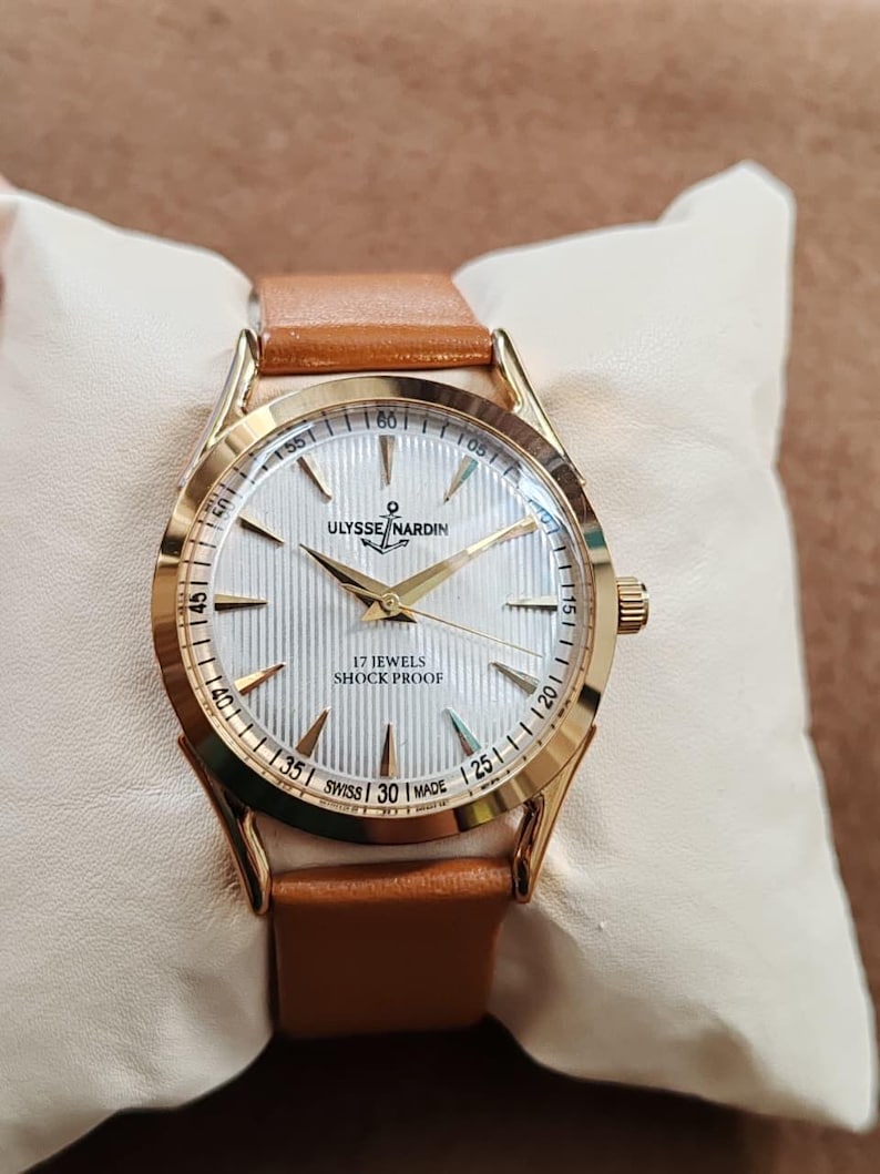 May include: A gold-tone wristwatch with a tan leather strap and a white dial featuring vertical pinstripes. The dial displays the text Ulysse Nardin with an anchor logo, 17 Jewels, Shock Proof, and Swiss Made, with gold-tone hands and markers.