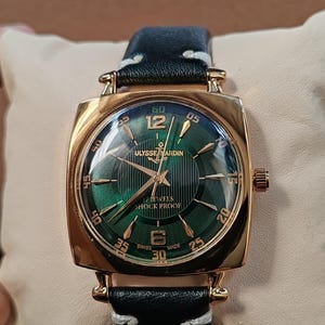 K&ouml;nnte beinhalten: Eine goldfarbene Armbanduhr mit quadratischem Geh&auml;use und gr&uuml;nem Zifferblatt mit Sonnenschliff, goldenen Indizes und Zeigern. Das Zifferblatt zeigt den Text ULYSSE NARDIN, 17 JEWELS, SHOCK PROOF und SWISS MADE. Sie hat ein schwarzes Lederarmband mit wei&szlig;en N&auml;hten.