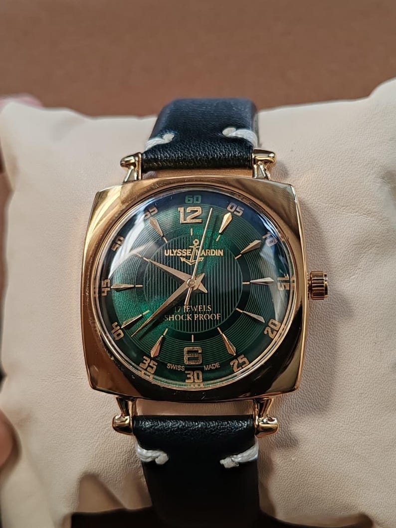 K&ouml;nnte beinhalten: Eine goldfarbene Armbanduhr mit quadratischem Geh&auml;use und gr&uuml;nem Zifferblatt mit Sonnenschliff, goldenen Indizes und Zeigern. Das Zifferblatt zeigt den Text ULYSSE NARDIN, 17 JEWELS, SHOCK PROOF und SWISS MADE. Sie hat ein schwarzes Lederarmband mit wei&szlig;en N&auml;hten.