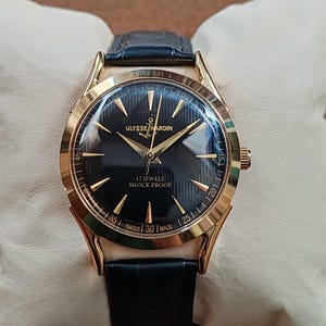 Puede incluir: Un reloj de pulsera dorado con esfera negra, manecillas e &iacute;ndices dorados. La esfera muestra el texto ULYSSE NARDIN, 17 JEWELS, SHOCK PROOF y SWISS MADE. Tiene una correa de cuero negro con acabado texturizado y una caja de forma redonda.