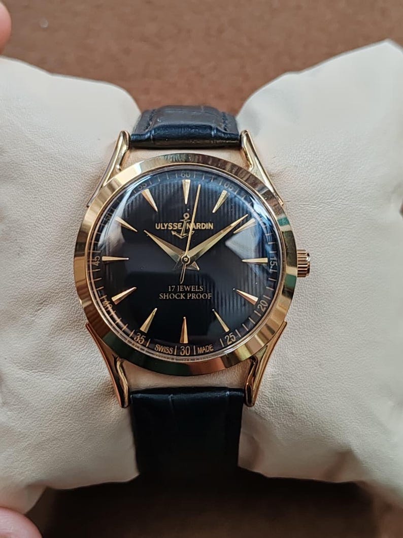 Puede incluir: Un reloj de pulsera dorado con esfera negra, manecillas e &iacute;ndices dorados. La esfera muestra el texto ULYSSE NARDIN, 17 JEWELS, SHOCK PROOF y SWISS MADE. Tiene una correa de cuero negro con acabado texturizado y una caja de forma redonda.