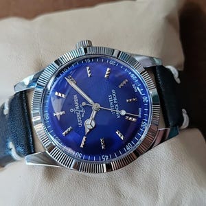 May include: A Ulysse Nardin wristwatch with a circular blue dial, silver-tone hands, and hour markers. The watch features a fluted silver-tone bezel, a black leather strap with white stitching, and text reading 17 JEWELS SHOCK PROOF SWISS MADE.