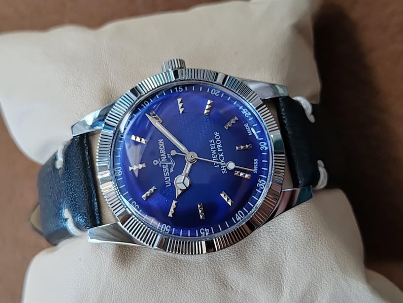 May include: A Ulysse Nardin wristwatch with a circular blue dial, silver-tone hands, and hour markers. The watch features a fluted silver-tone bezel, a black leather strap with white stitching, and text reading 17 JEWELS SHOCK PROOF SWISS MADE.
