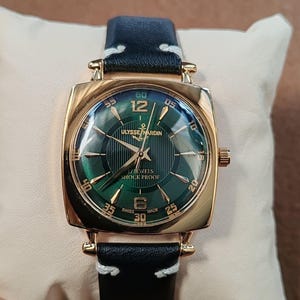 K&ouml;nnte beinhalten: Eine goldfarbene Armbanduhr mit quadratischem Geh&auml;use und dunkelgr&uuml;nem Zifferblatt mit Sonnenschliff, goldenen Indizes und Zeigern. Das Zifferblatt zeigt den Text ULYSSE NARDIN, 17 JEWELS, SHOCK PROOF und SWISS MADE. Sie hat ein schwarzes Lederarmband mit wei&szlig;en N&auml;hten.